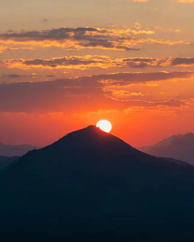 The sun rising directly behind a dark mountain peak, spilling gold and orange light across a layered, cloud-streaked sky, with hazy blue ridgelines fading into the distance.